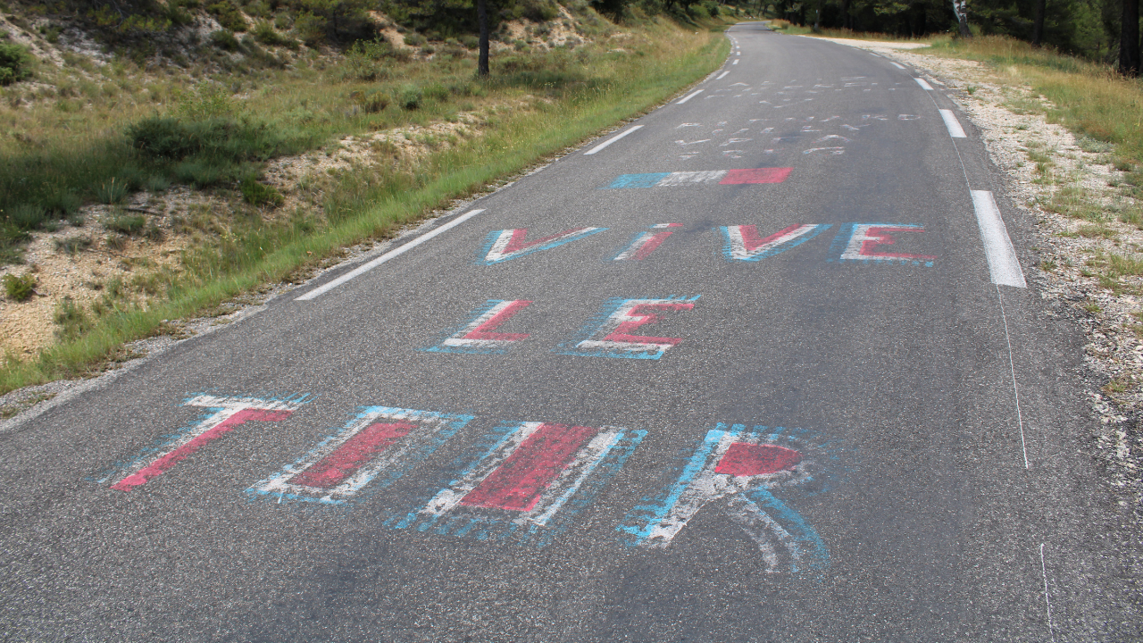 The phrase "VIVE LE TOUR" painted in large, colorful letters on the asphalt of a road, likely in celebration of the Tour de France cycling race.