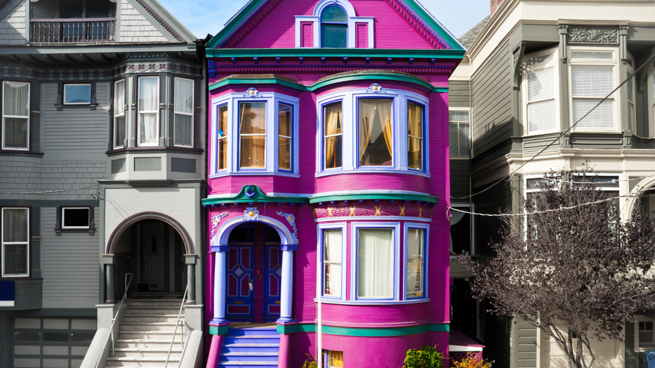 A vibrant purple and pink Victorian house with ornate detailing, nestled between more subdued colored homes.