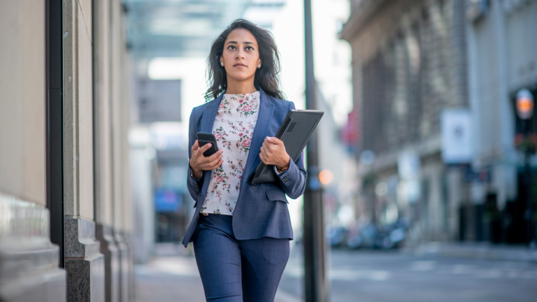A young professional woman in a business suit walks down an urban city street holding a phone in one hand and a laptop in the opposite arm