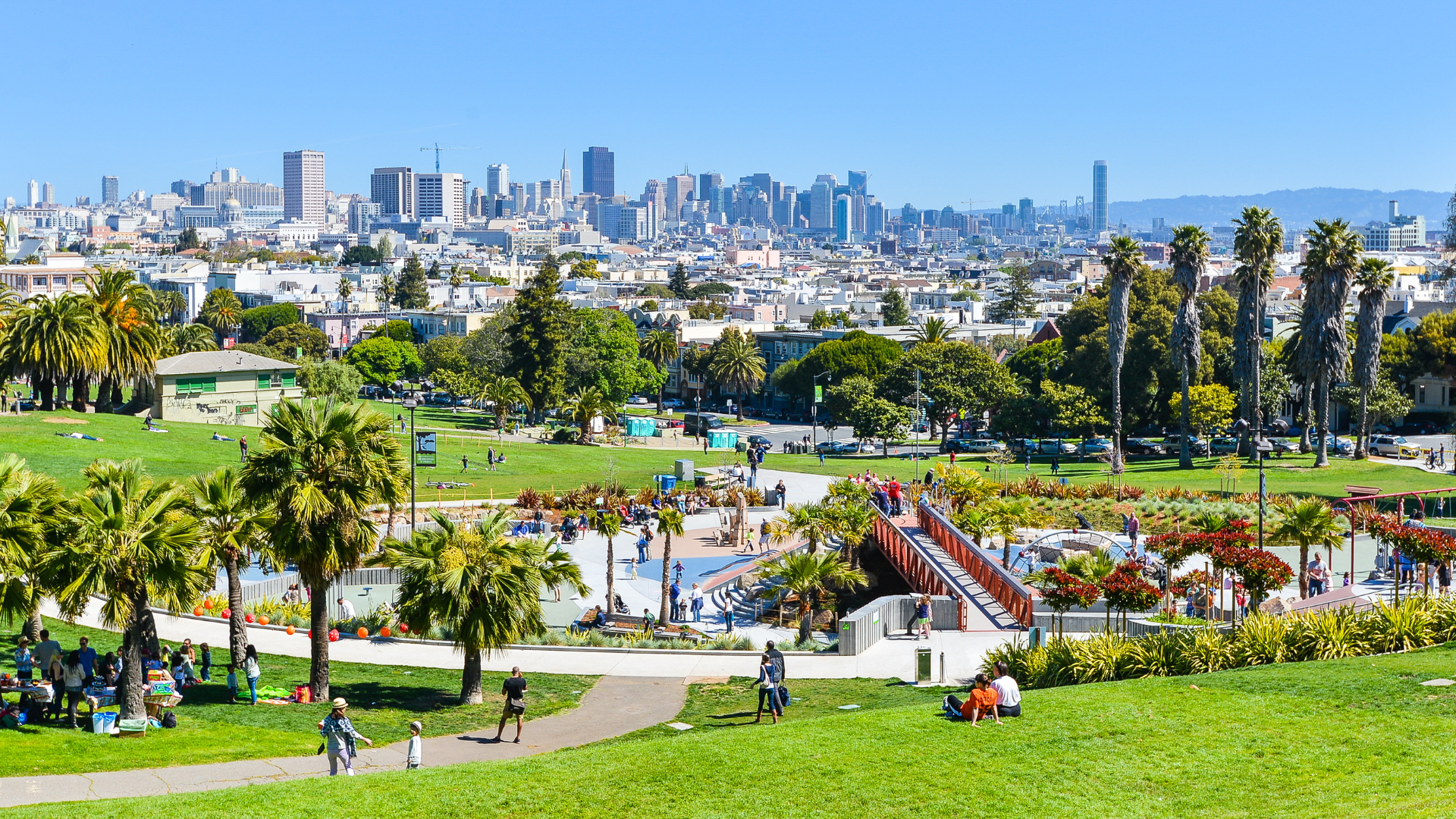 A vibrant urban park with green lawns and palm trees set against the backdrop of the San Francisco skyline. People are scattered across the grass, some relaxing on blankets while others walk along the paths or gather in small groups. In the center is a playground area and a bridge-like structure connecting different sections of the park. The tall buildings of San Francisco's downtown area rise in the distance under a clear blue sky, adding contrast to the greenery in the foreground. The atmosphere is lively and sunny, suggesting a pleasant day outdoors.